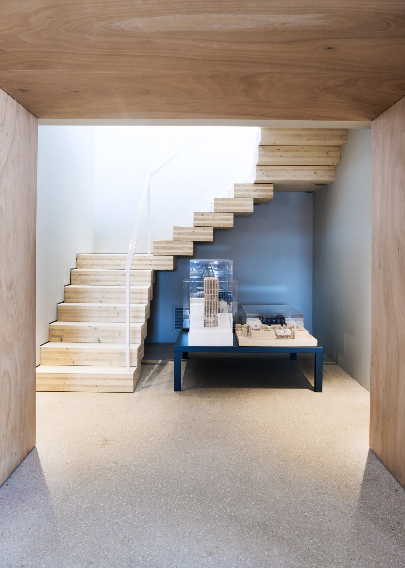 Wooden staircase with light wood steps ascending against white walls and blue-grey concrete wall in modern office space.