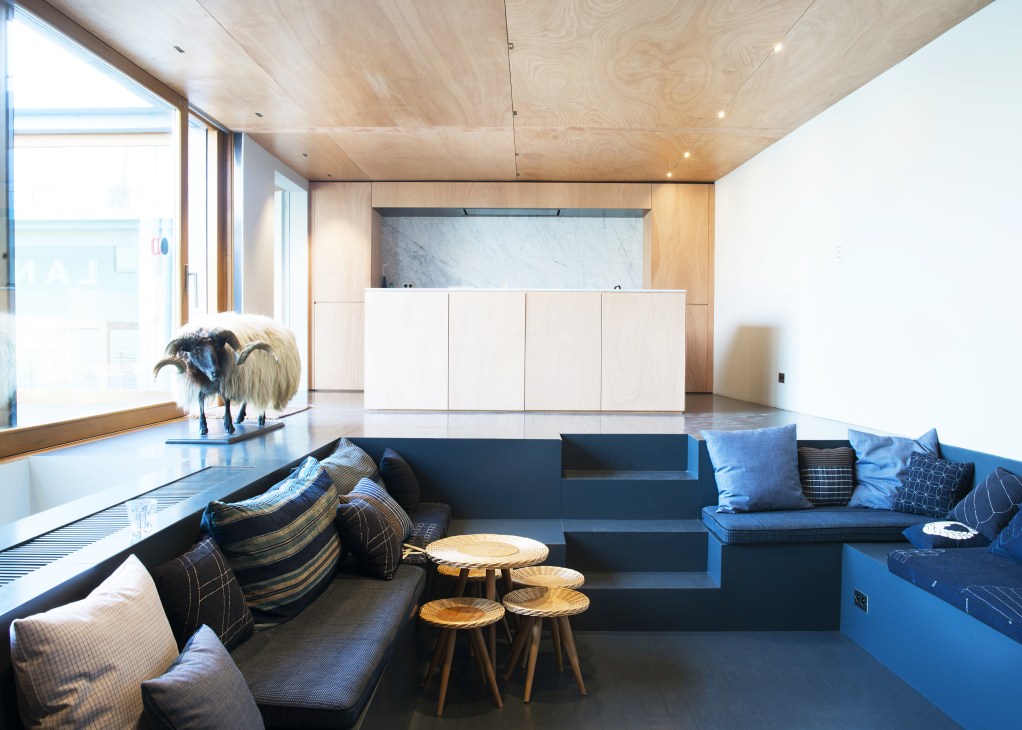 Modern recreational space with blue seating, wooden accents, and a sculptural goat figurine near large windows.