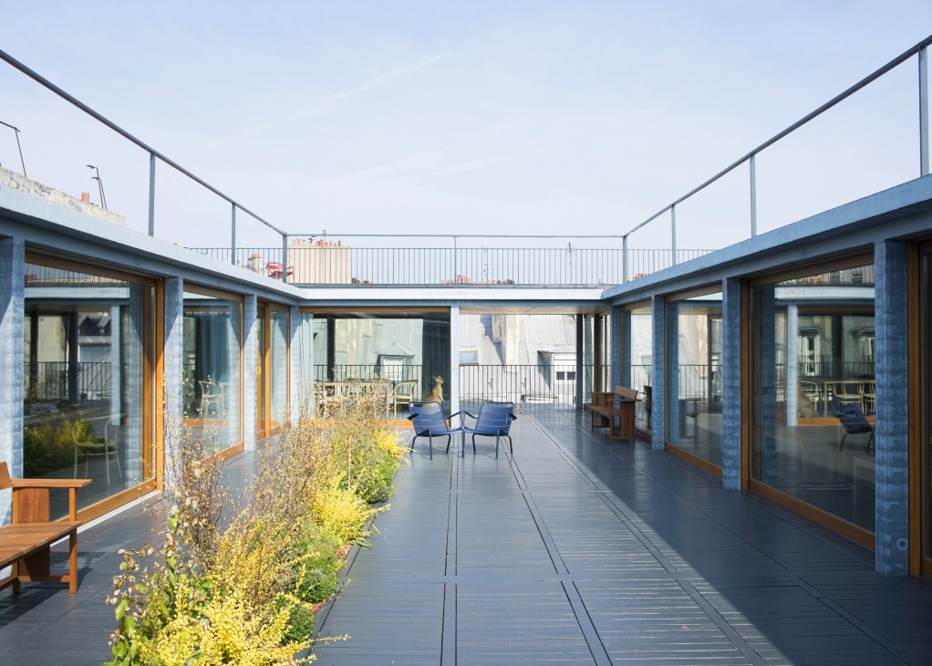 Modern rooftop courtyard with blue-grey concrete pillars, wooden decking, climbing ivy, and contemporary seating areas surrounded by glass-fronted offices.