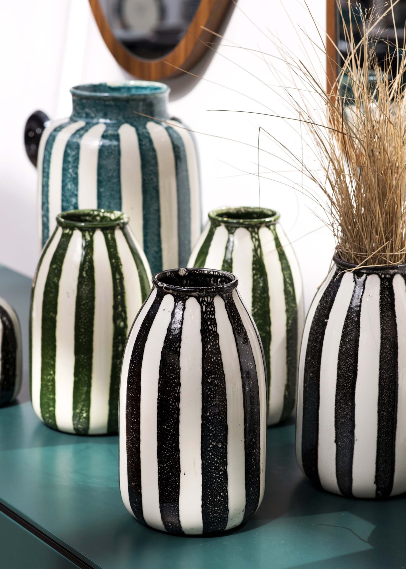 Striped ceramic vases in black, green and white arranged with dried grasses and a wooden mirror.
