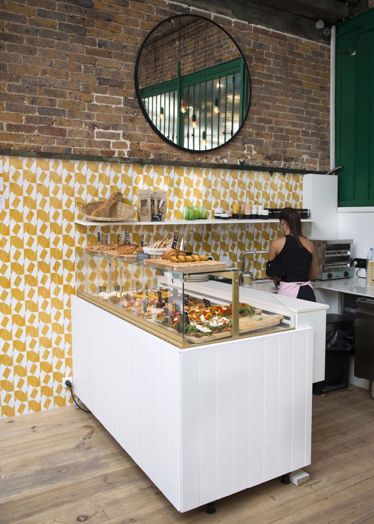 Patisserie counter with yellow geometric tile backsplash, round mirror, and pastry display cases.