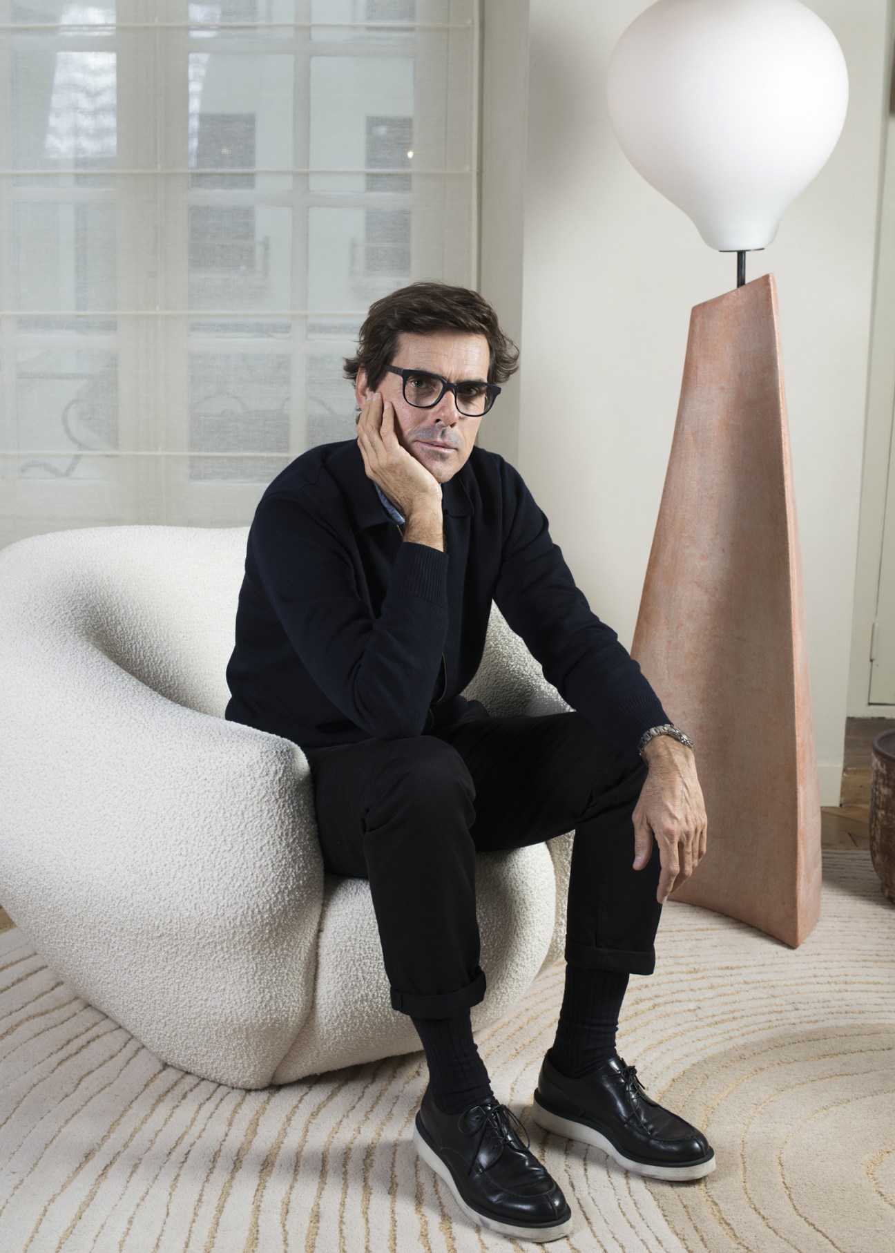 Man in black clothing sitting on white sculptural chair next to modern floor lamp in minimalist studio space.
