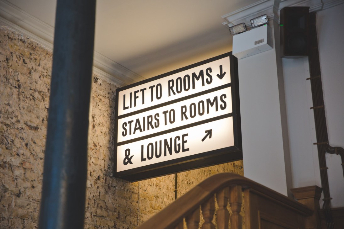 Illuminated directional sign reading 'LIFT TO ROOMS, STAIRS TO ROOMS & LOUNGE' mounted on textured brick wall