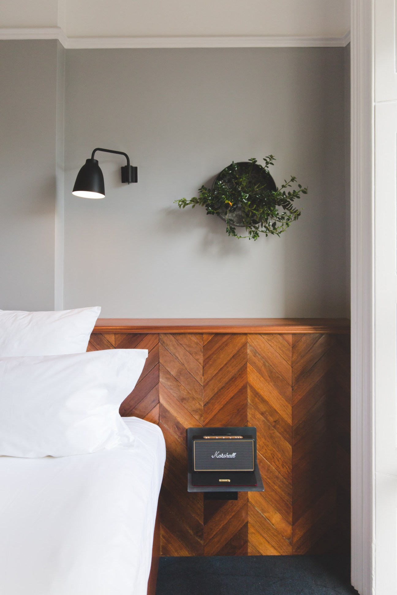 Hotel bedroom with wooden chevron sideboard, black wall lamp, potted plant, and white bedding with Marshall amplifier on nightstand.