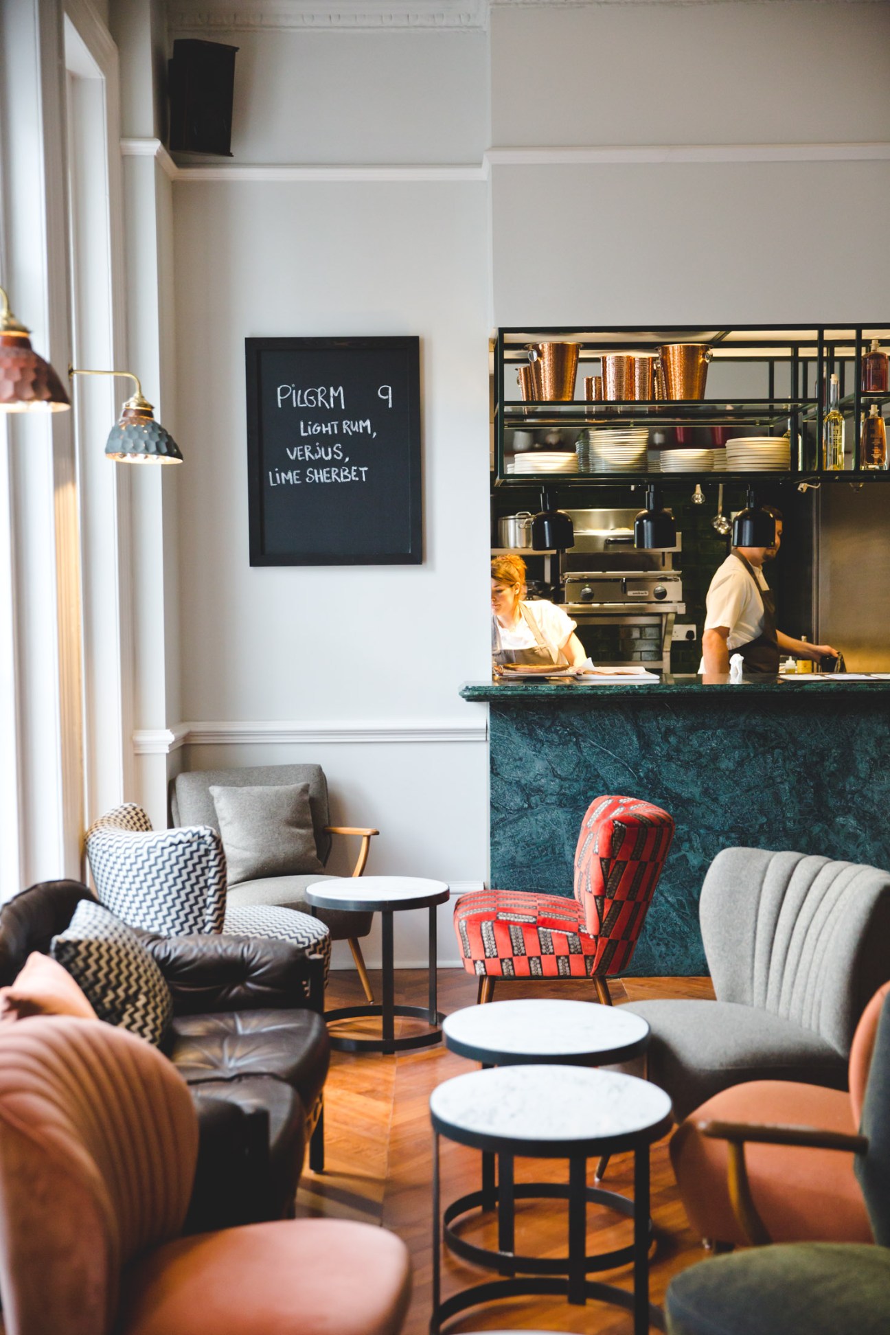 The Pilgrm hotel bar with teal-green counter, staff working behind, chalkboard menu, and seating area with patterned chairs.