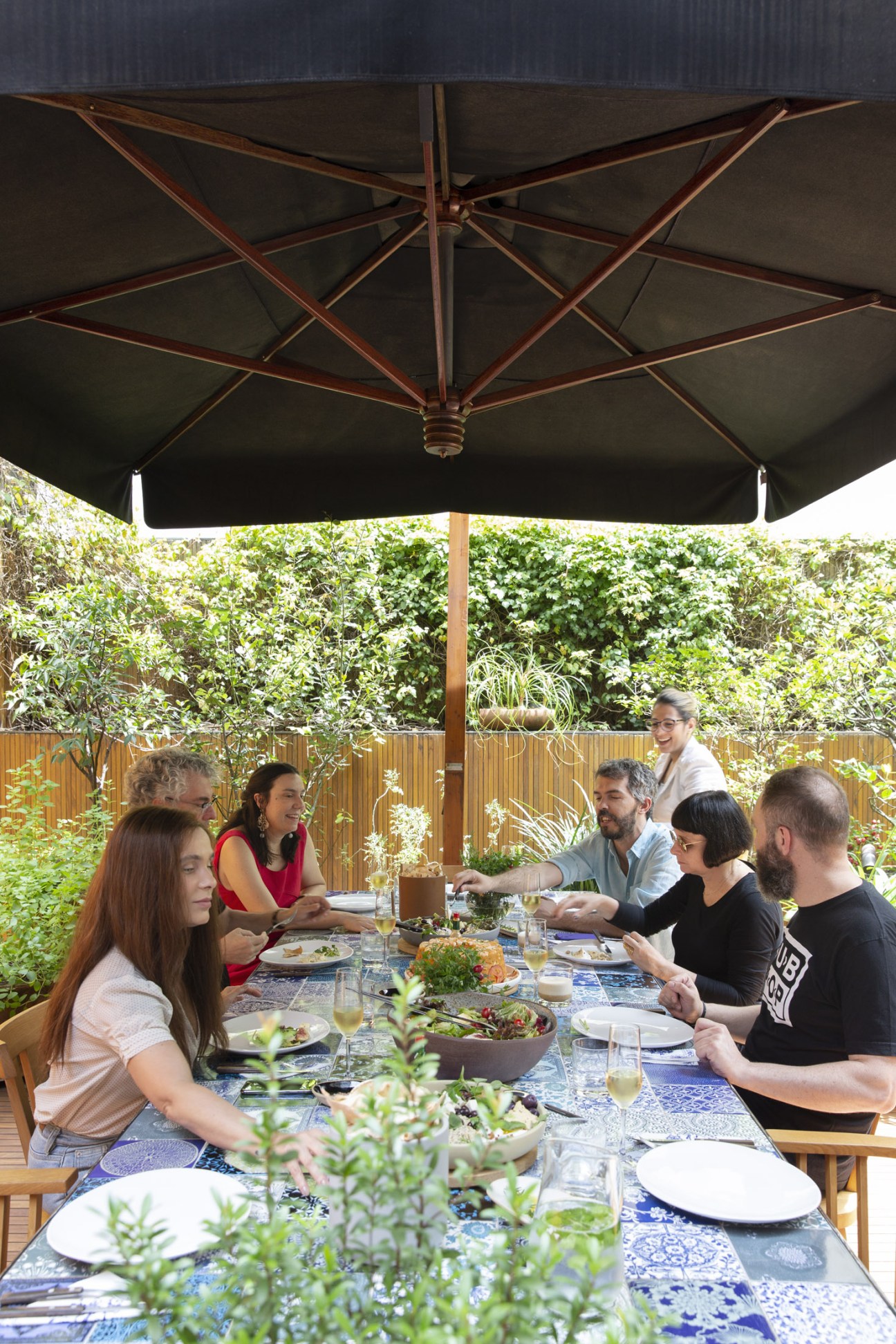 People dining outdoors on a patio under a black umbrella surrounded by lush greenery at Studio mk27