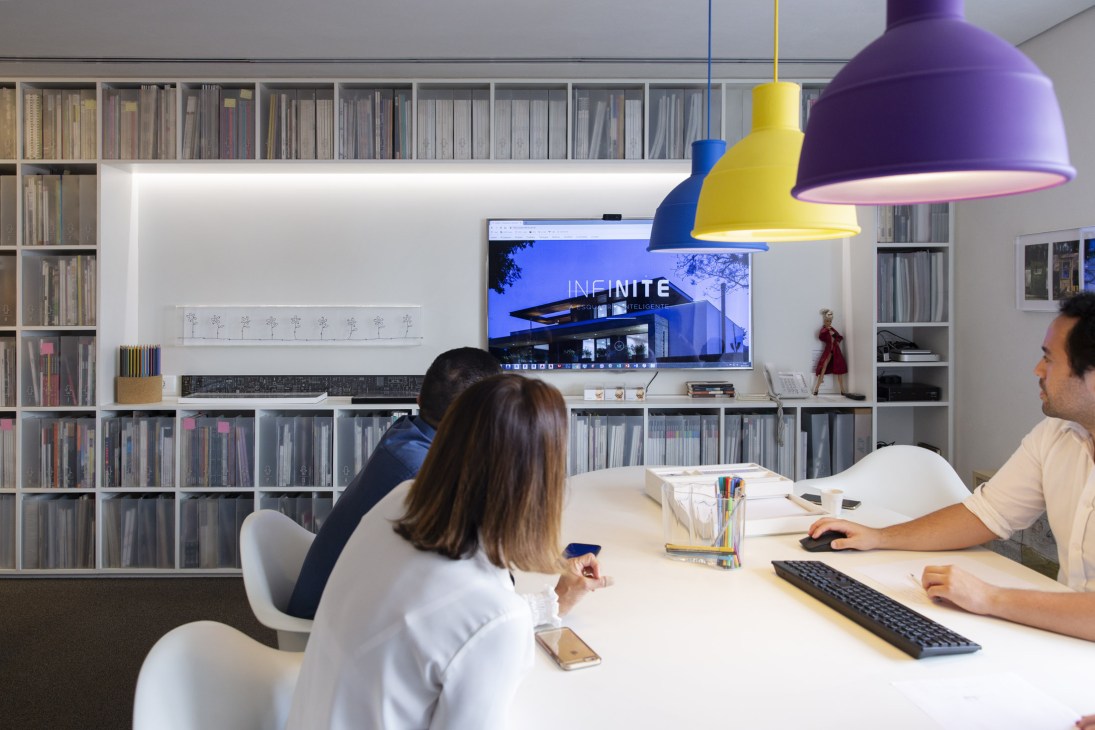 Two architects working at a desk in Studio mk27's open-plan office with shelving and a digital display.