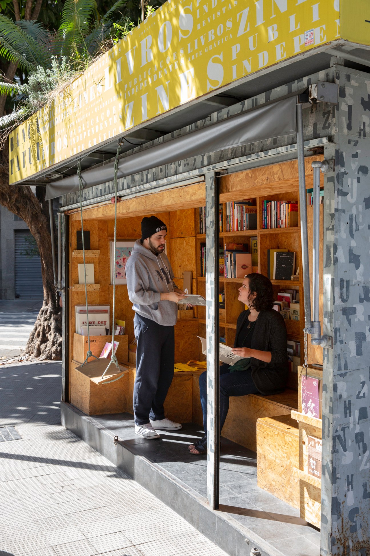 Two people at Banca Tatuí newsstand, one standing and one sitting, with books and publications visible inside the kiosk