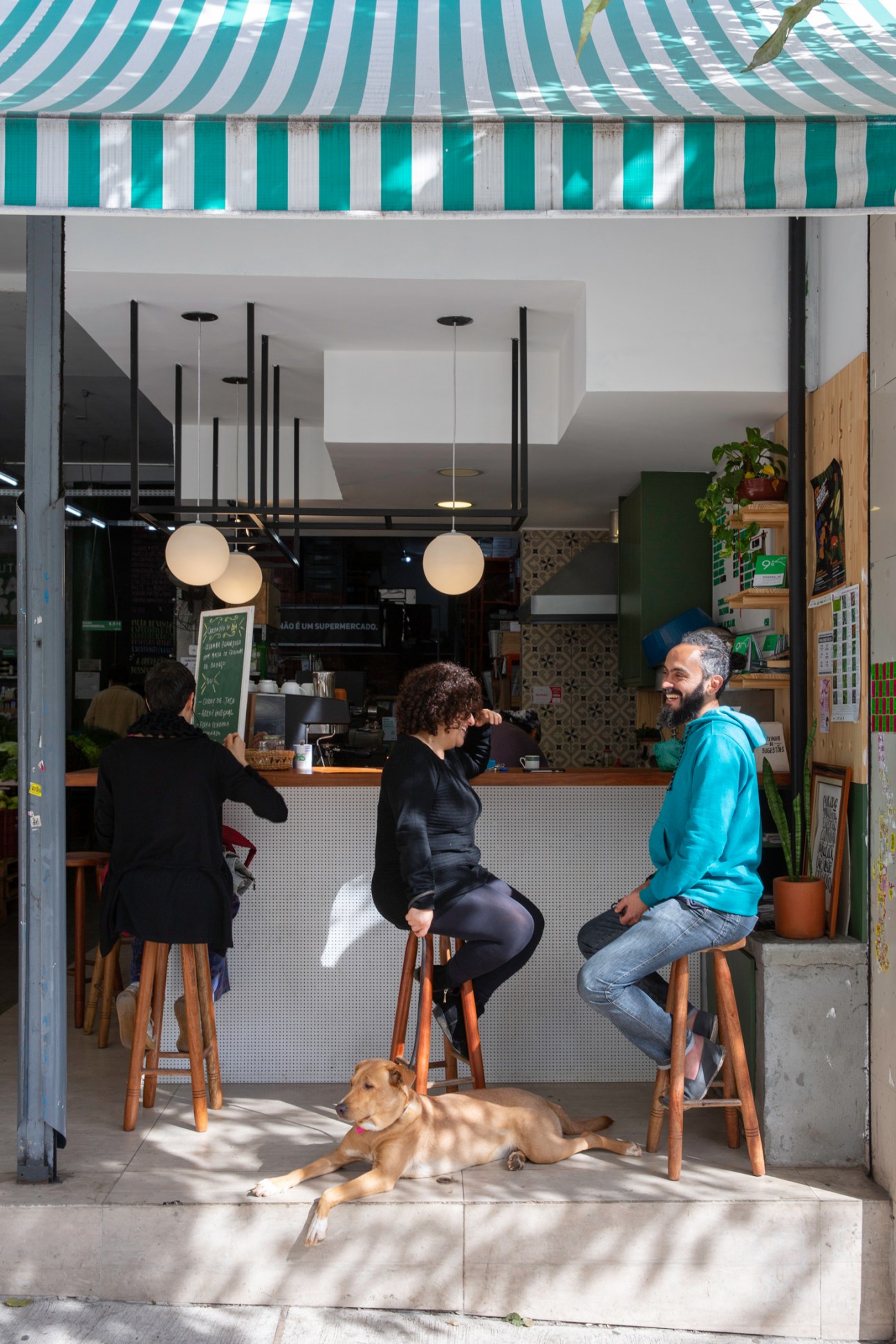 People sitting at outdoor counter of cafe with green striped awning, dog lying on sidewalk below