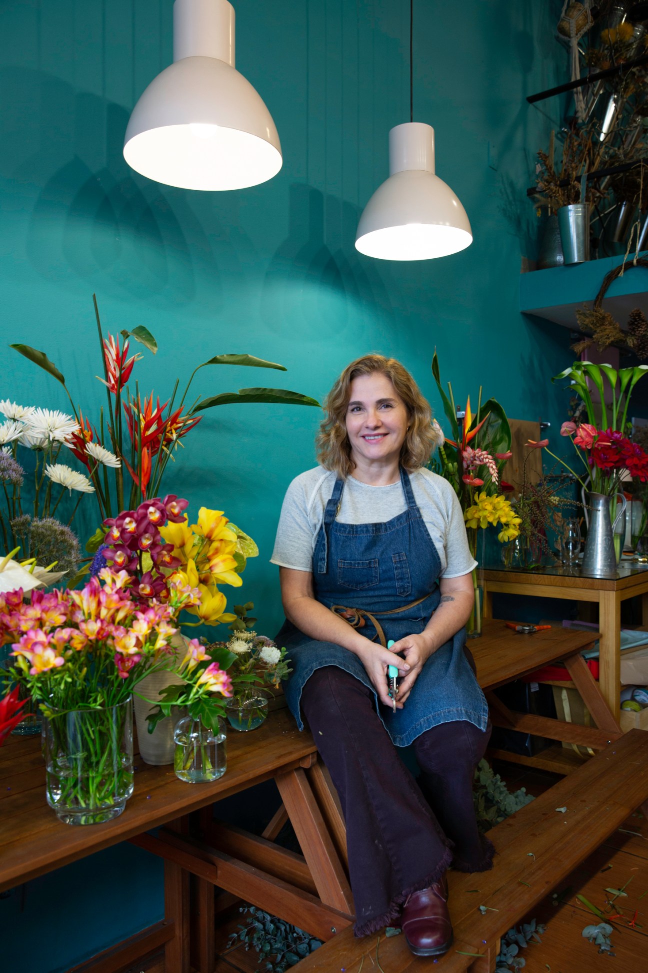 Woman in denim apron sitting on wooden bench in flower shop surrounded by colorful tropical arrangements