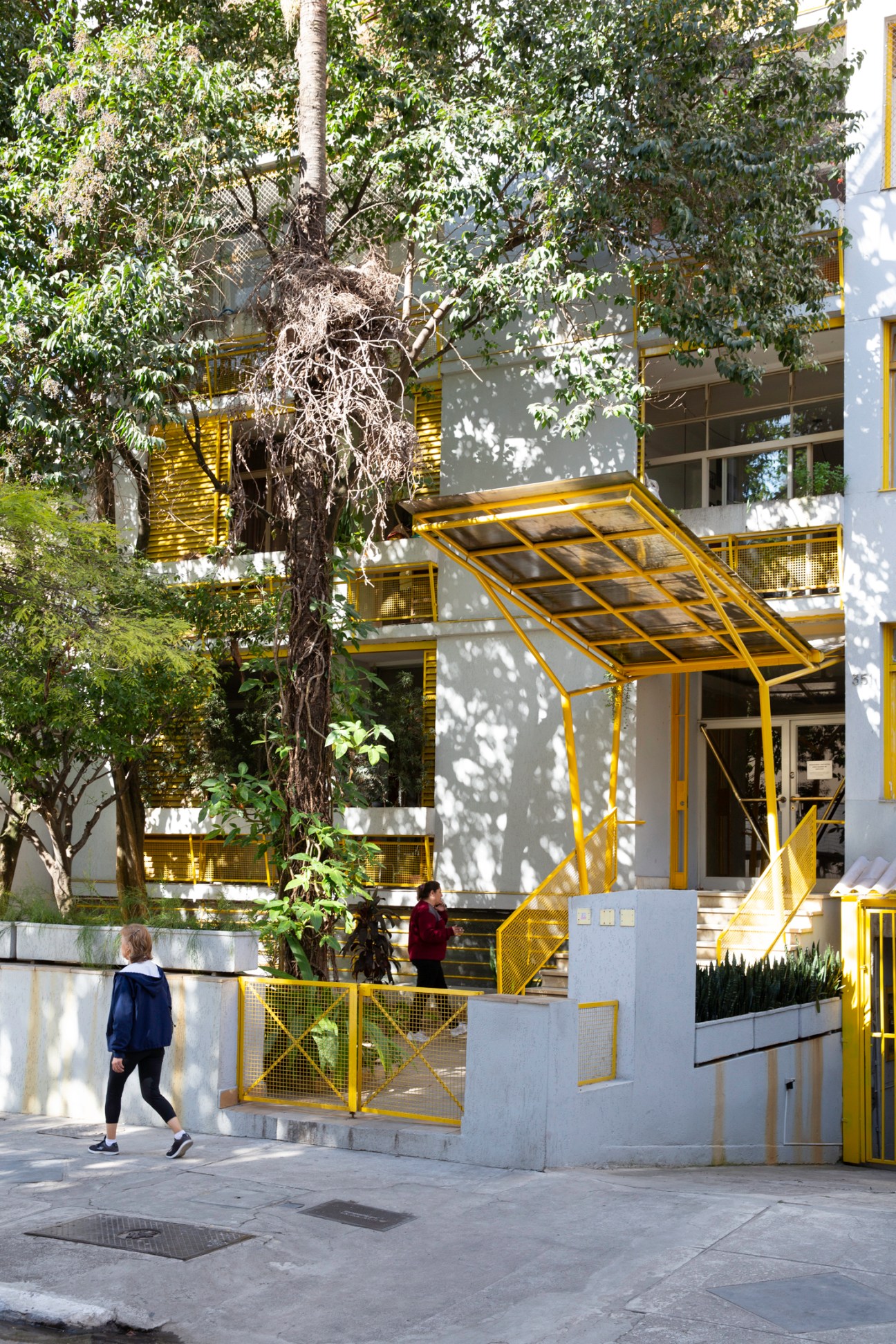 Modern residential building with yellow metal architectural elements, balconies, and trees casting shadows
