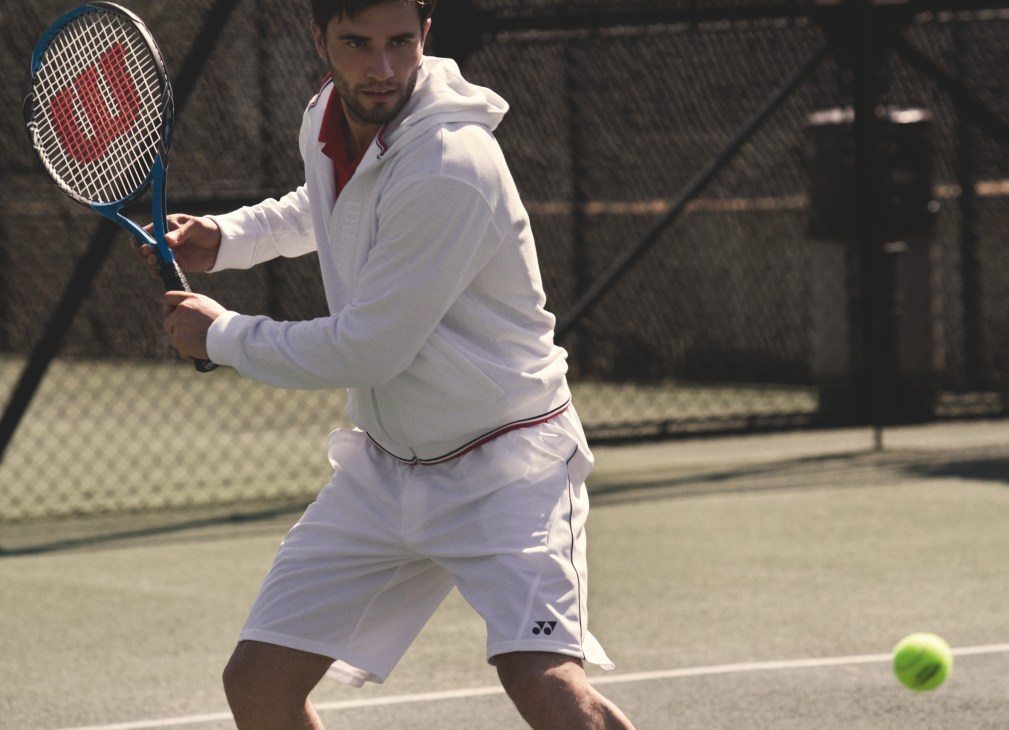 Male model in white Wilson jacket and Yonex polo shirt and shorts holding Wilson racquet on tennis court.