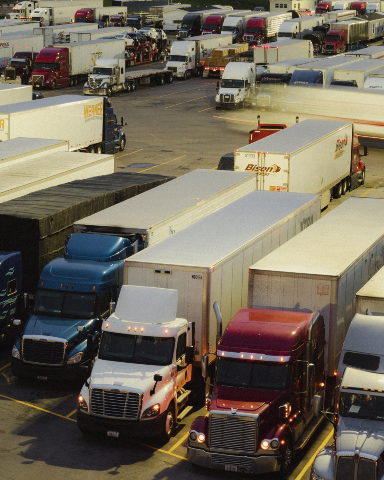 Aerial view of a large truckstop parking lot filled with semi-trucks and trailers at dawn or dusk.