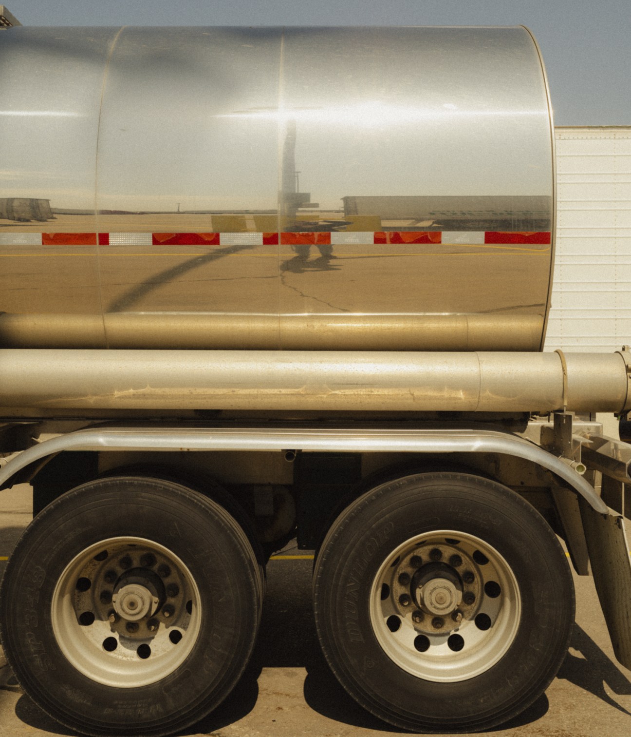 Large tanker truck with reflective polished surface parked at a truckstop, showing a driver's shadow on the side.