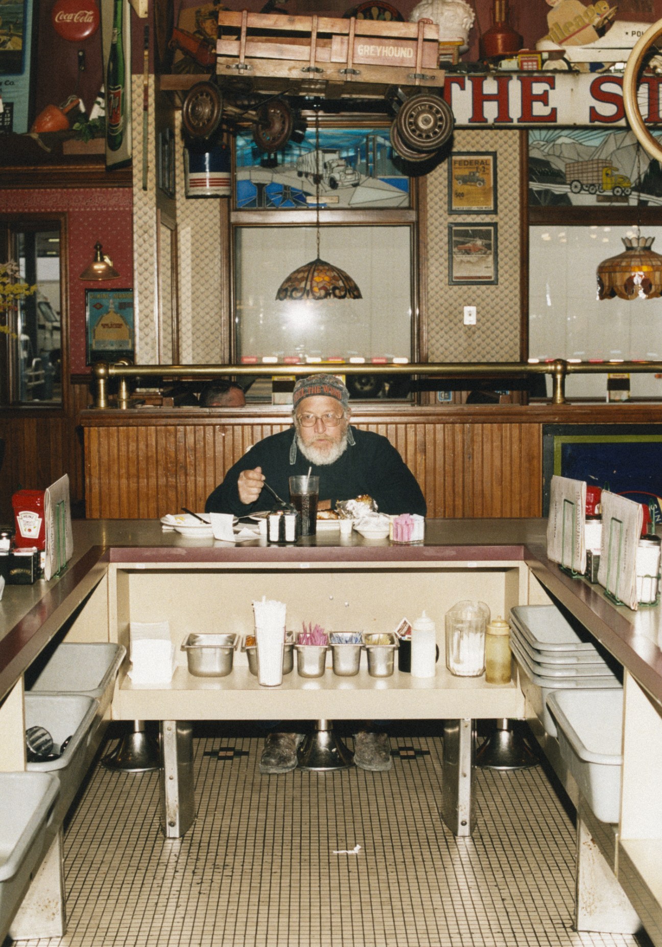 An older man in glasses and a cap sits at a vintage diner counter eating, surrounded by classic Americana décor and vintage truck memorabilia.