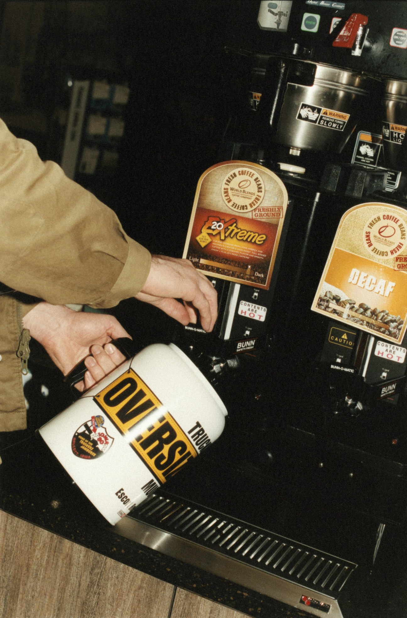A trucker's hand pouring coffee from a carafe into a white mug at a truckstop.