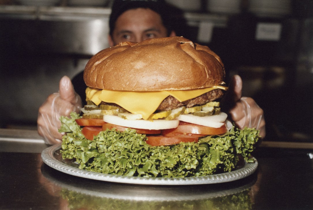 A person holds up R Place's four-pound Premium Ethyl hamburger with cheese, lettuce, tomato, and onion on a plate.