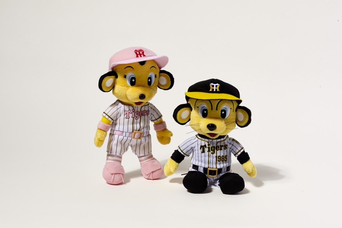 Two plush tiger mascot toys dressed in Hanshin Tigers baseball uniforms with pink and black caps.