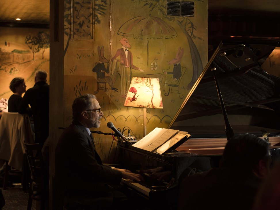 Pianist performing at grand piano in Bemelmans Bar with golden lighting and colorful 1940s murals by Ludwig Bemelmans visible on walls.