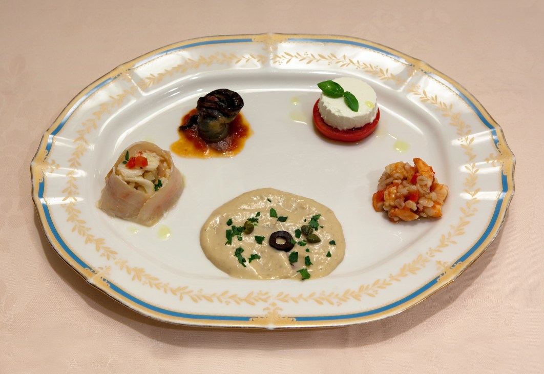 An ornate white plate with blue and gold trim displaying five Italian appetizers including bruschetta, mozzarella, and seafood preparations.