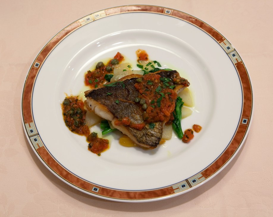 Plated isaki fish with tomato sauce, capers, and green vegetables on white porcelain.