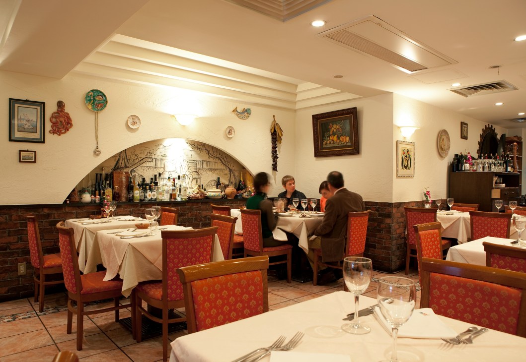 Interior of Elio Locanda Italiana restaurant in Tokyo with red upholstered chairs, white tablecloths, and Italian décor on walls.