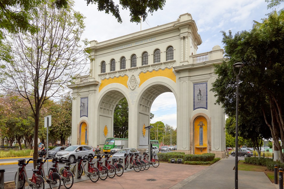 White neoclassical arches with yellow accents frame a busy street intersection with bike sharing stations in foreground