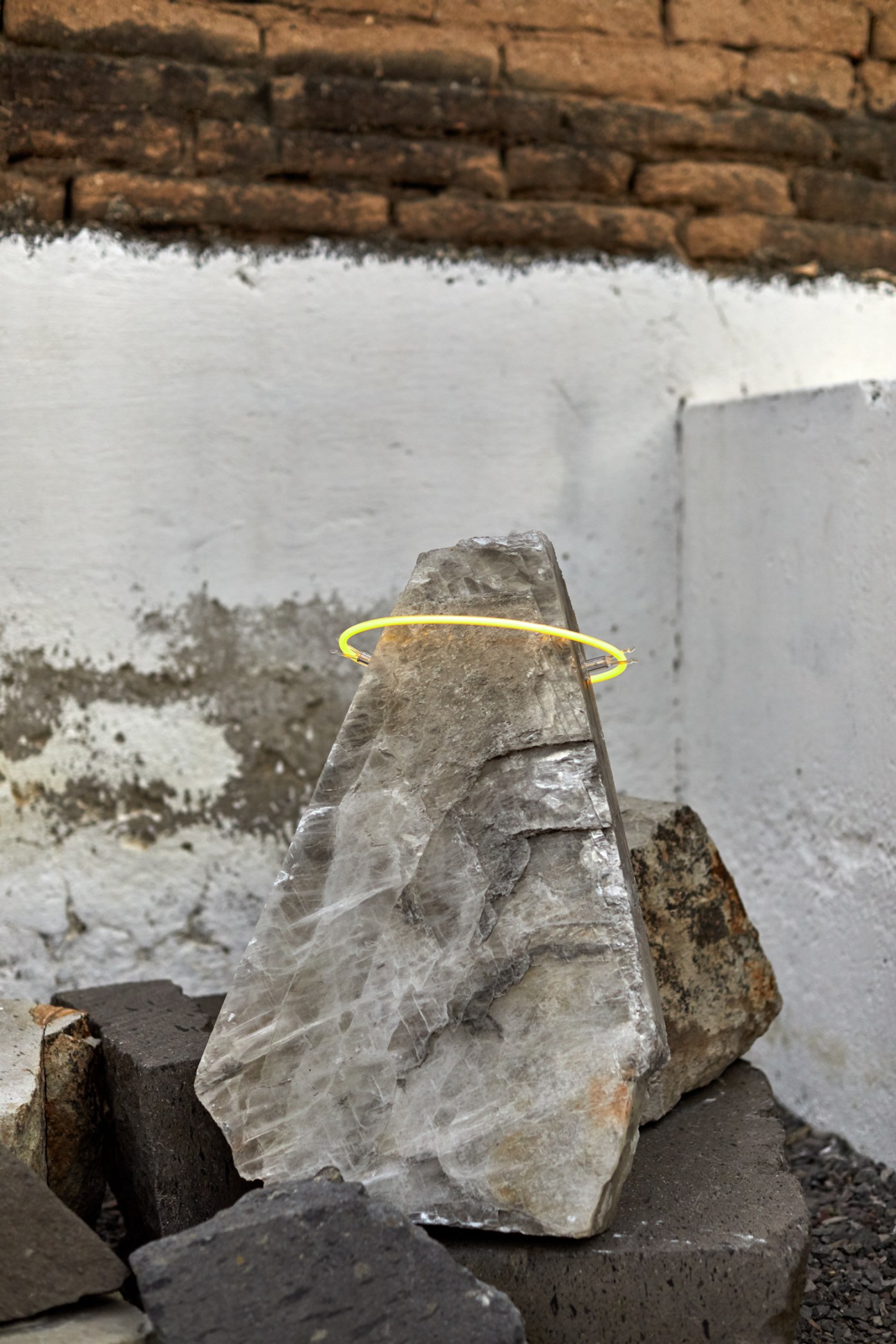 Stone sculpture with yellow neon ring around middle against weathered brick and white walls