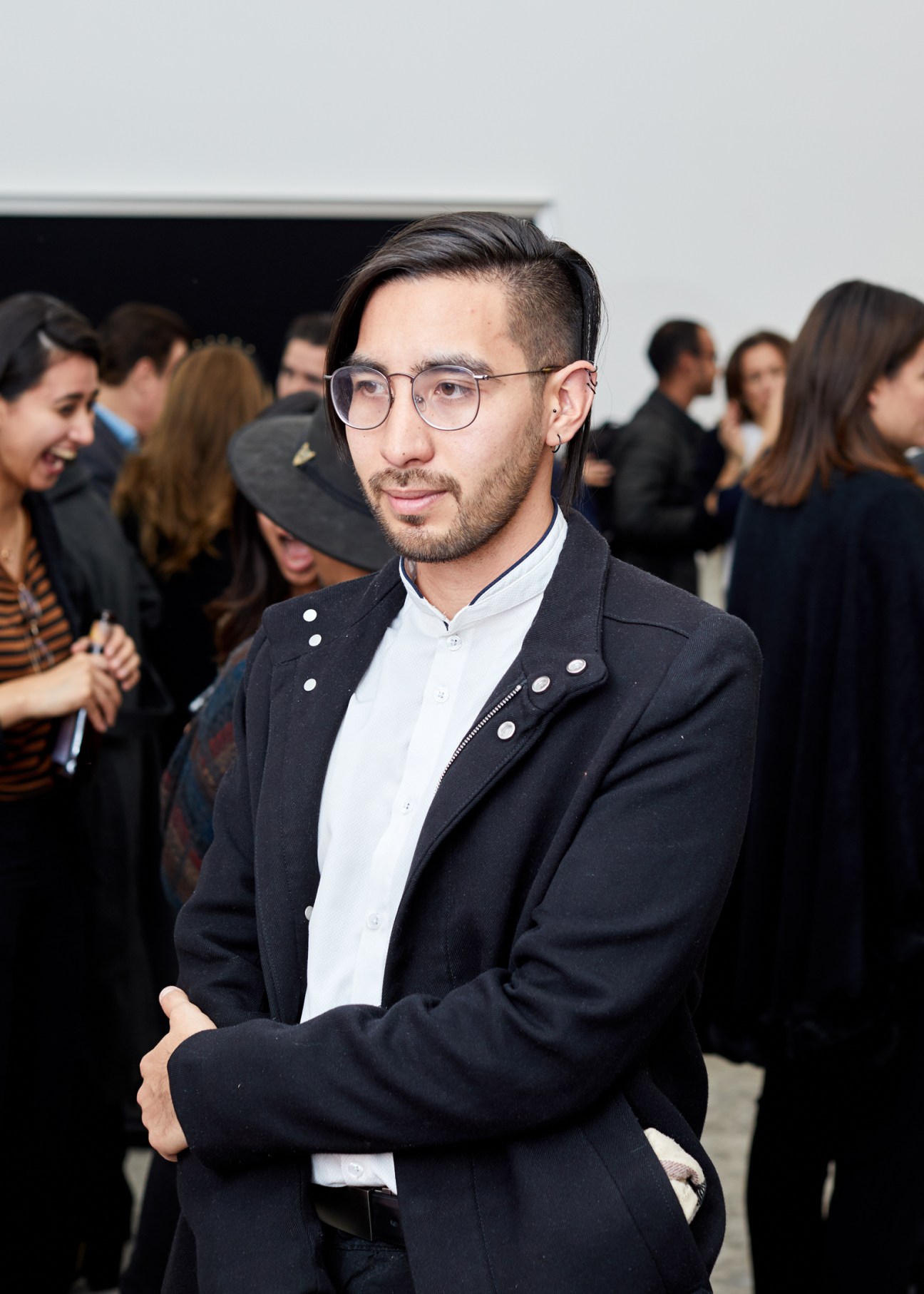 Young man with glasses and beard in dark blazer at art gallery opening with attendees in background