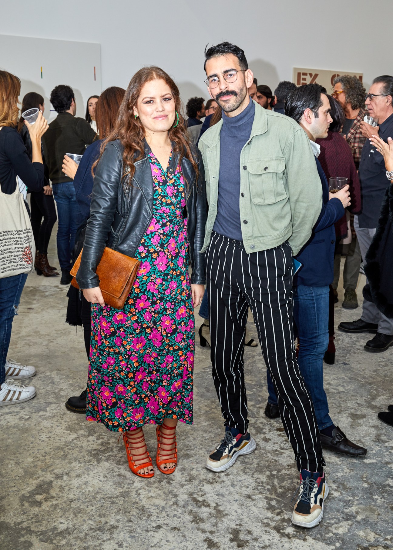 Woman in floral dress and leather jacket with man in striped pants at art gallery opening with crowd in background