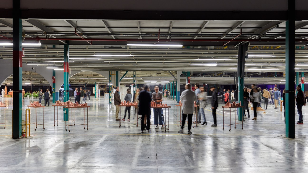 People viewing art installations on wheeled display tables in a large industrial space with exposed ceiling