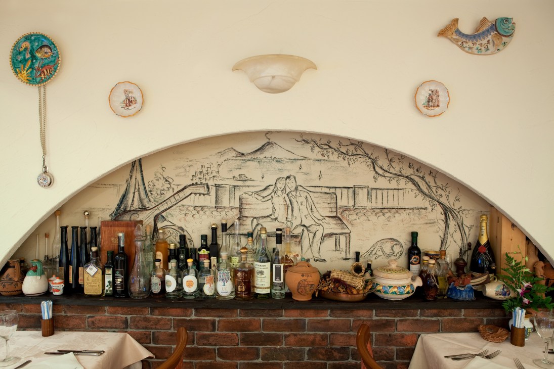 Elio's restaurant wall display featuring Italian ceramics, decorative plates, fish ornaments, and bottles of wine and spirits arranged on a brick ledge beneath a large mural.