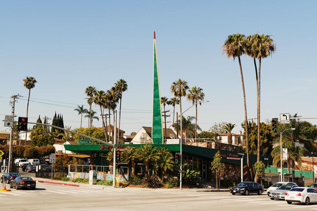 Green Googie-style diner building with pointed spire, palm trees, and street view in Los Angeles.