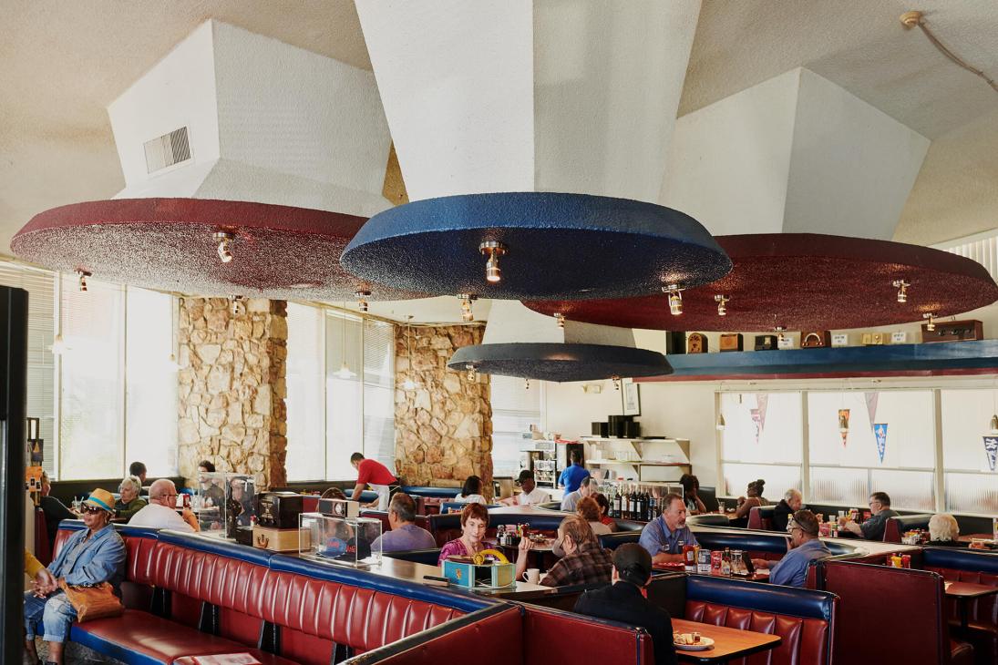 Interior of Dinah's diner showing circular UFO-shaped ceiling fixtures in red, blue, and brown suspended above red vinyl booths with diners seated throughout.