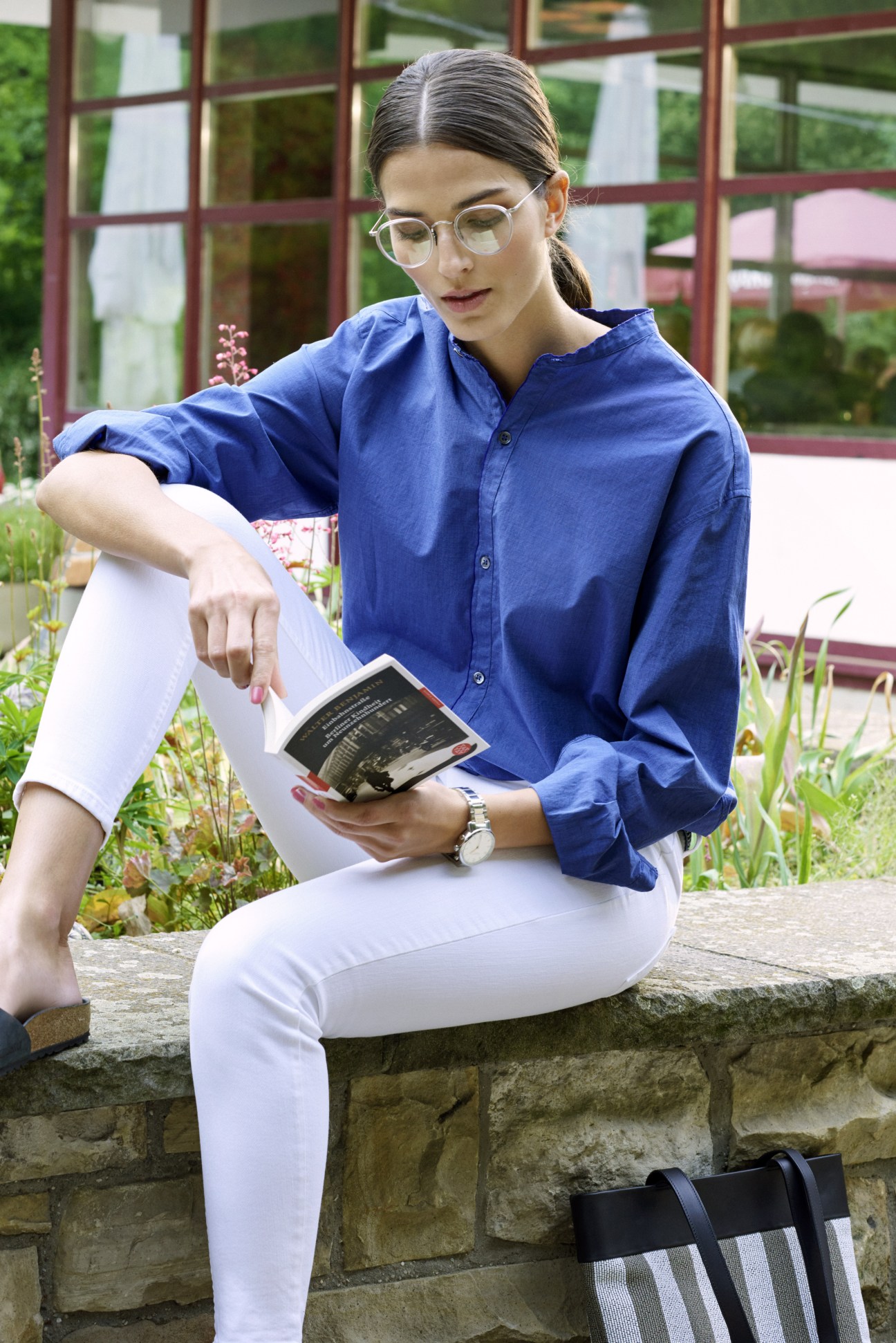 Woman in blue shirt and white trousers reading a book while seated outdoors by a garden.