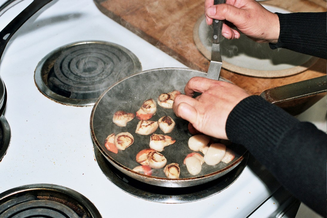 Gregory pan-frying scallops in a dark skillet on a stovetop, adding raw scallops with one hand