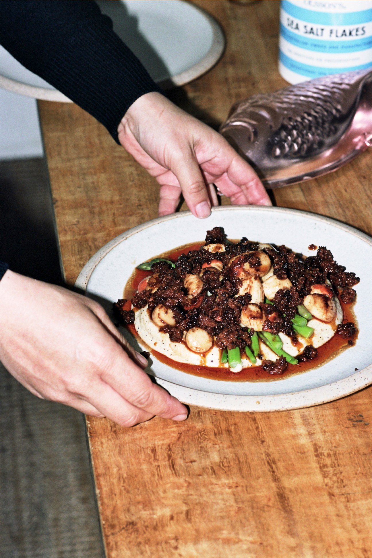 Hands presenting a plated dish with scallops, buckwheat polenta, and dark garnish on white ceramic plate