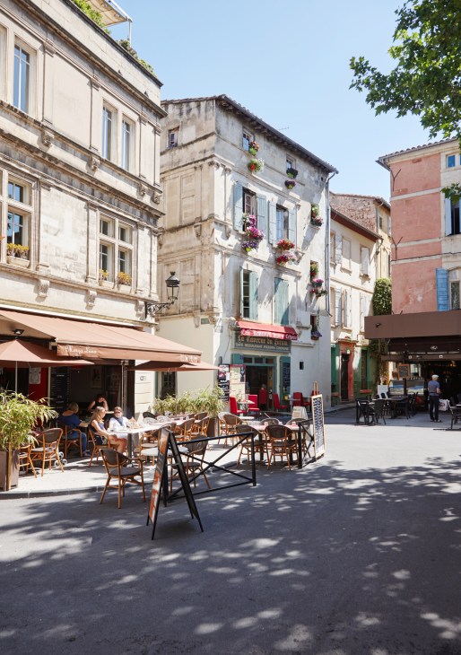 A sunny plaza in Arles with historic stone buildings, outdoor café seating, and flowering window boxes.