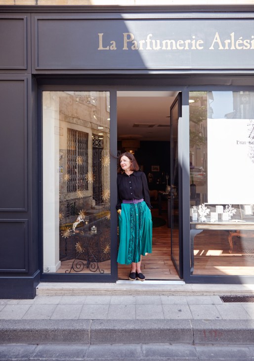 Woman standing in doorway of La Parfumerie Arlésienne boutique in Arles wearing a black shirt and teal skirt.