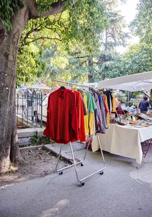 Colorful clothing displayed on racks at an outdoor Saturday market in Arles under a large tree.
