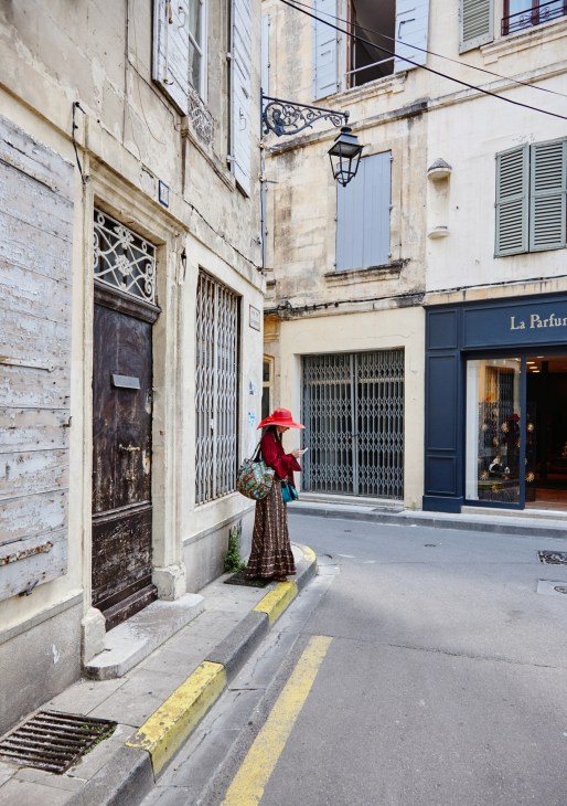 Woman in red hat and patterned dress walking through a narrow Arles street past historic buildings and La Parfumerie Arlésienne boutique.