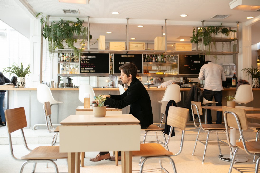 Modern café interior with wooden chairs, white tables, and a customer seated at a table with pendant lamps overhead.