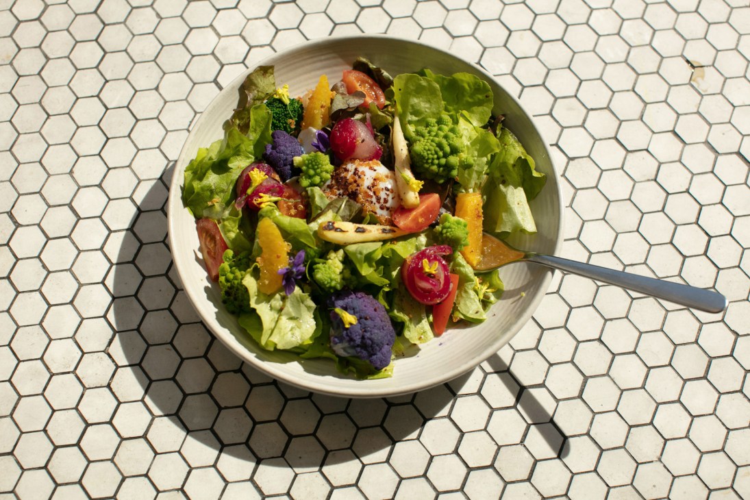 A colorful salad with mixed greens, purple cauliflower, yellow peppers, red tomatoes, and edible flowers on a white plate.