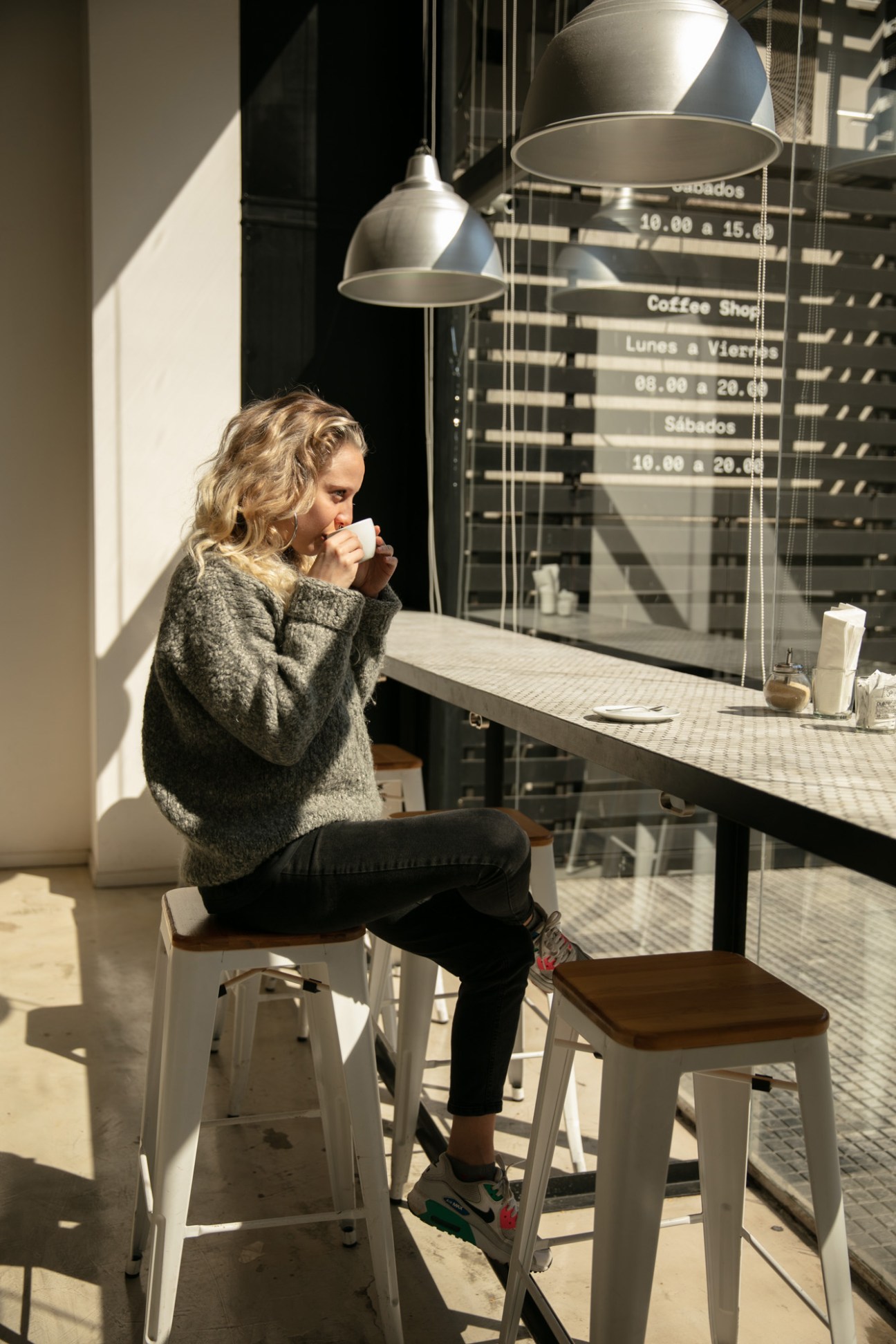 Woman sipping coffee at a minimalist café with industrial pendant lamps and window signage for Lab Tostadores de Café.