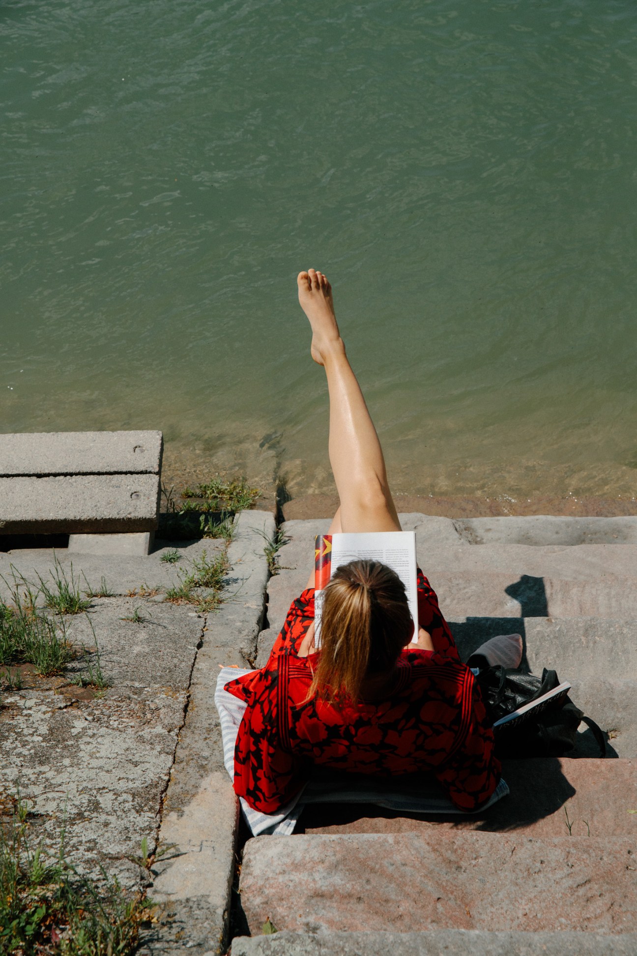 Person in red patterned shirt reading by the Rhine with one leg stretched up toward the water.