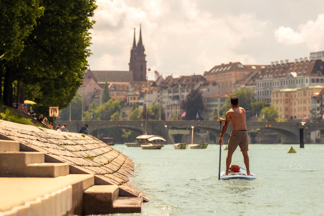 Person stand-up paddleboarding on the Rhine River with Basel's cathedral and bridges visible in the background.