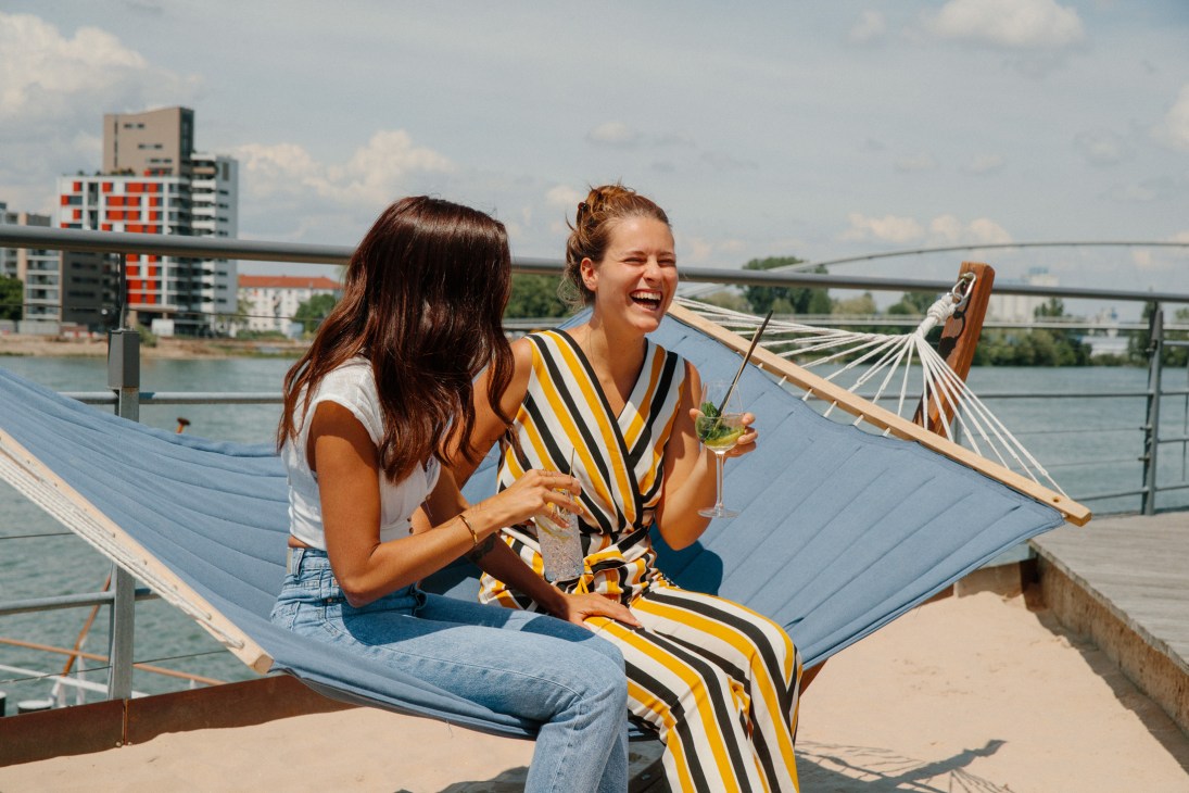 Two women relaxing in a hammock by the Rhine in Basel, holding drinks with the city skyline and bridges in the background.