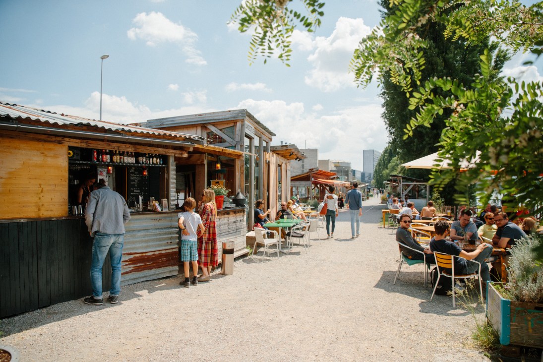 Wooden beachside kiosks and dining area along the Rhine with people seated at tables and standing at a counter.