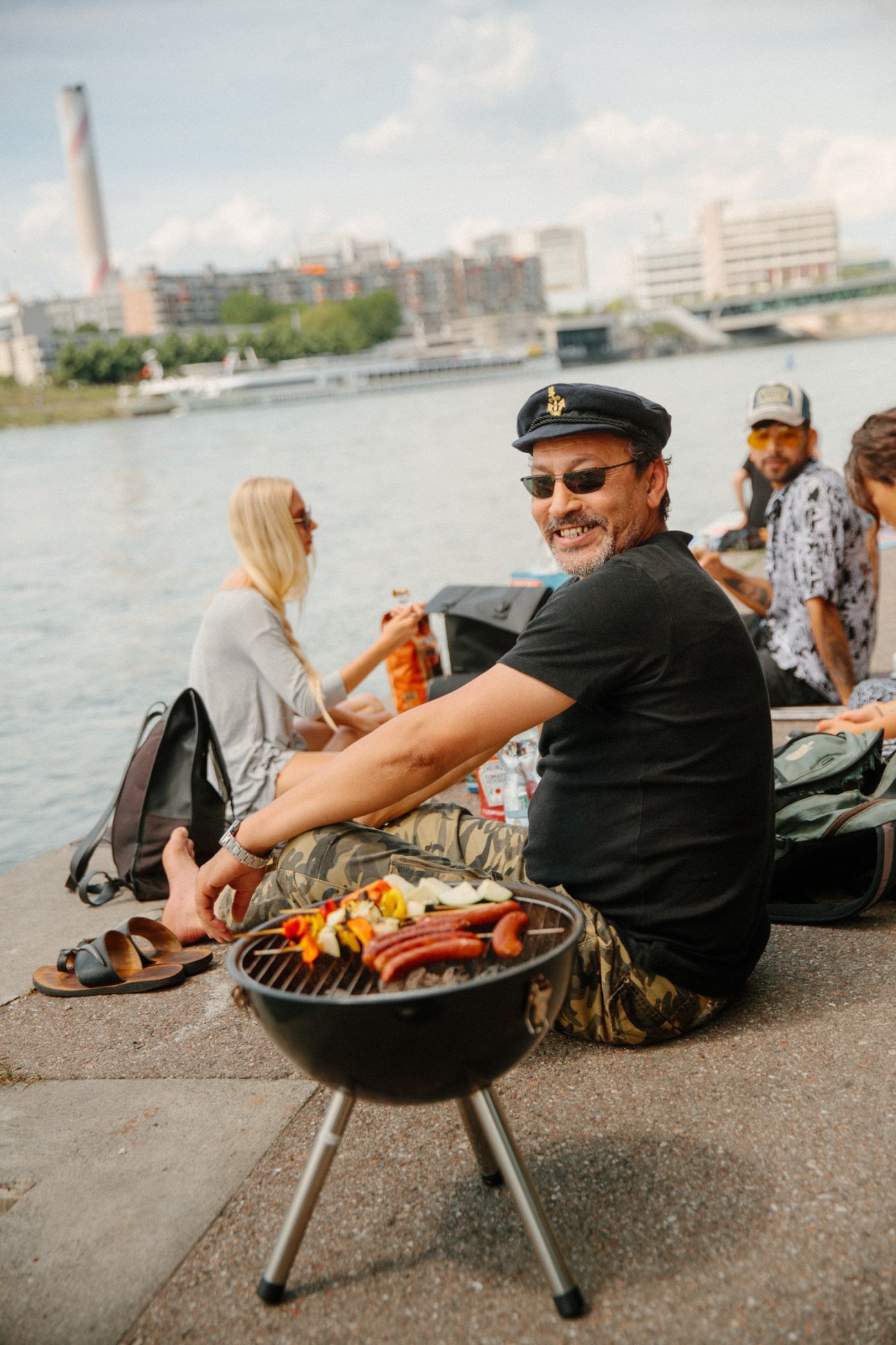A man in a captain's hat grilling food by the Rhine river in Basel with people and waterfront buildings behind him.