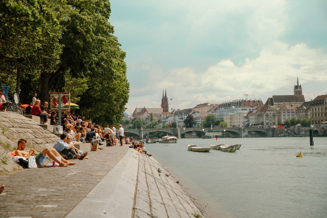 People sunbathing on concrete steps along the Rhine riverbank in Basel with the Mittlere Brücke bridge and historic architecture visible across the water.